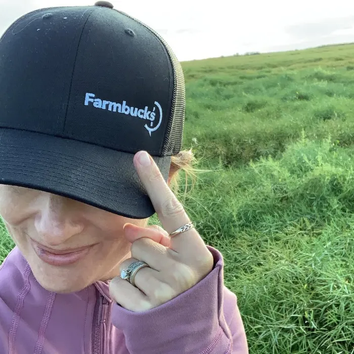 Lynn Dargis in the field wearing a Farmbucks hat on her head pointing to Farmbucks logo on her hat with her finger and a smile on her face.