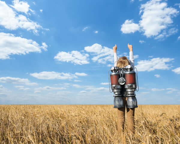 A boy standing in a field with a jet-pack on his back and his arms in the air.